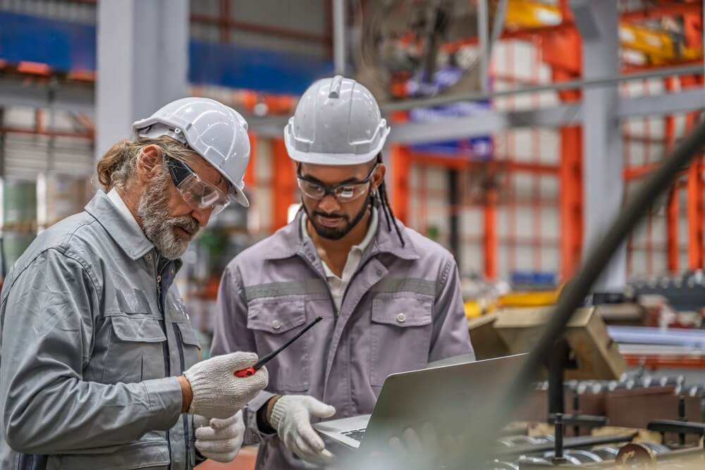 Two men, one silver-haired and another with braids, both wearing grey hazmat suits, protective glasses, and hard hats while examining an item.